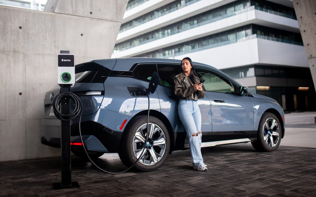 A woman stands beside a gray BMW iX electric vehicle connected to a SWTCH charging station, holding a phone